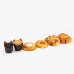 An Assortment of 6 Mini Breakfast Pastries, 2 Muffins, 2 Danish, 1 Croissant and 1 Straight Chocolate Croissant on a light grey background