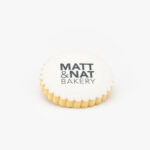 A Round Celebration Logo Cookie with a Matt & Nat Bakery Logo on a light grey background