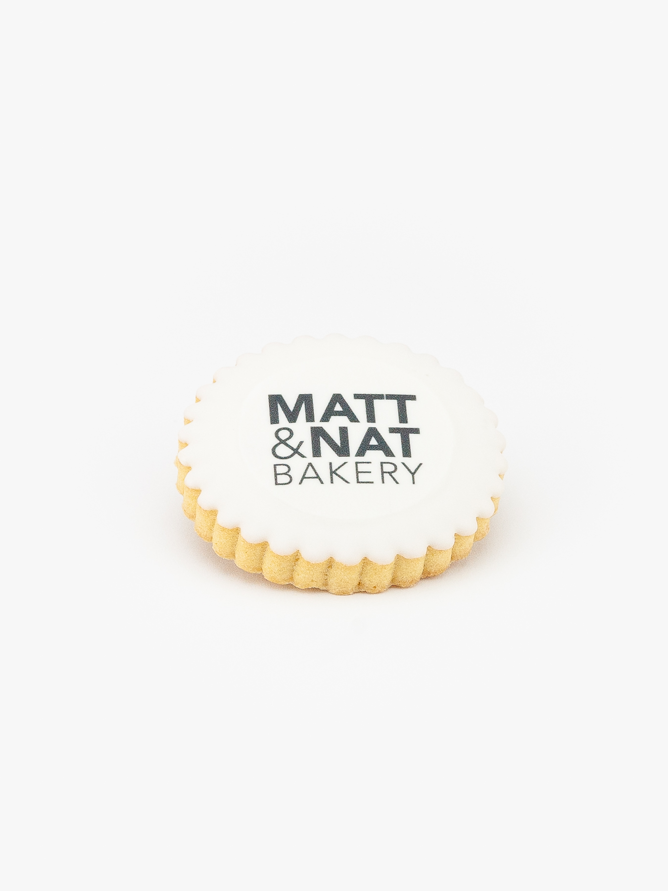 A Round Celebration Logo Cookie with a Matt & Nat Bakery Logo on a light grey background